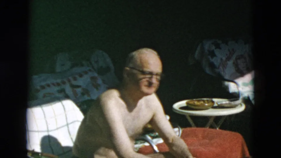 Shirtless older man with glasses sits in bright outdoor light beside a towel-covered chair and a small table with food or drink items.