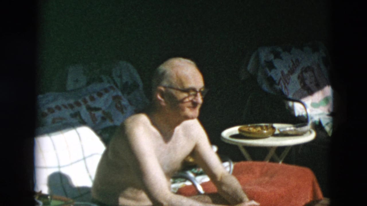 The man smiles or talks while seated outdoors, with a small table, towels, and shaded greenery behind him.