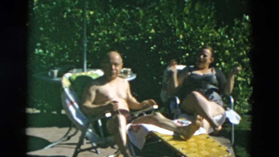 Two adults relax in folding lawn chairs in sunlight, with towels, a small table, and dense greenery around the seating area.