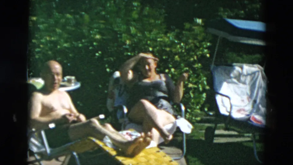 The seated pair relax outdoors as the woman adjusts her hair, surrounded by lawn chairs, towels, greenery, and strong sunlight.
