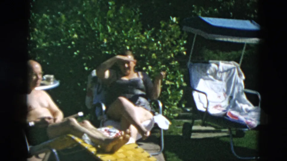 Adults relax in folding chairs and a lounge chair outdoors, with dense greenery and dappled sun around them.