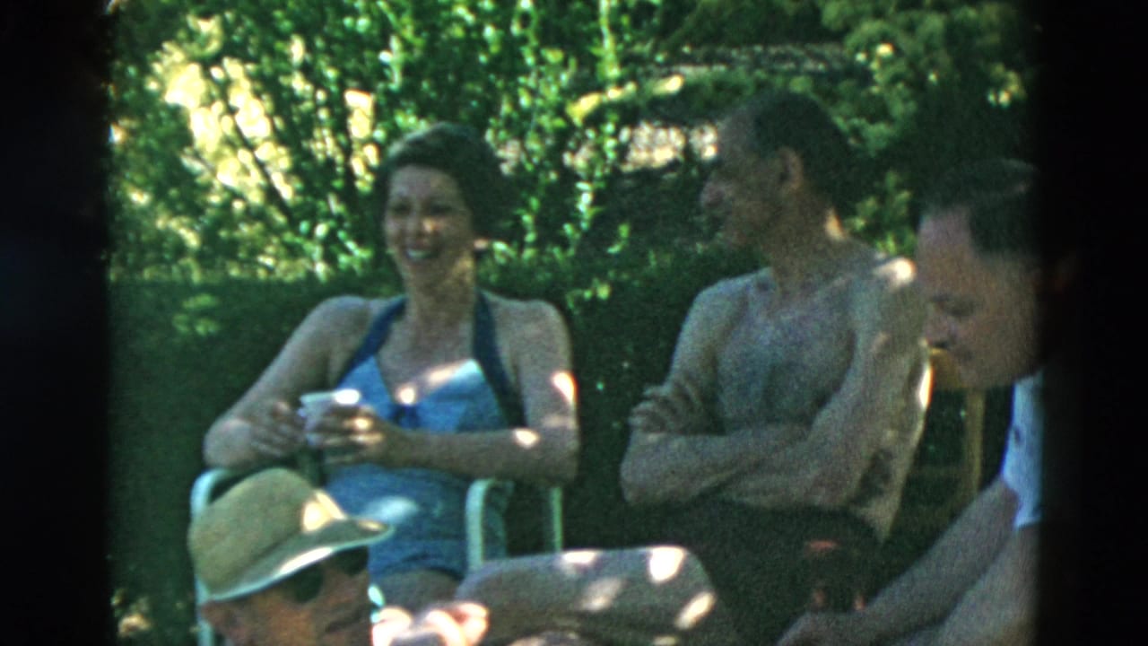 A woman in a blue one-piece swimsuit smiles while seated near a shirtless man and another adult in a garden setting.