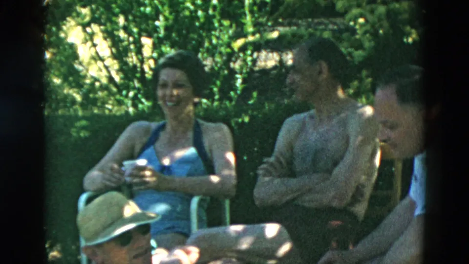 A woman in a blue one-piece swimsuit smiles while seated near a shirtless man and another adult in a garden setting.