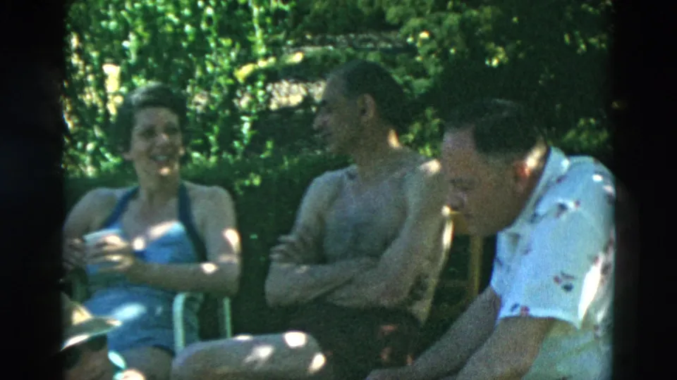 A woman in a swimsuit and two men sit in lawn chairs, with one man leaning forward during a casual conversation.