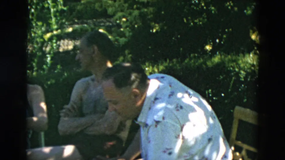 A shirtless seated man and a man in a patterned white shirt appear in conversation beside lawn chairs and garden greenery.
