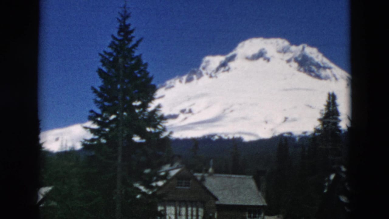 A snow-capped mountain rises beyond evergreen trees and a rustic building roofline in archival home-movie footage.