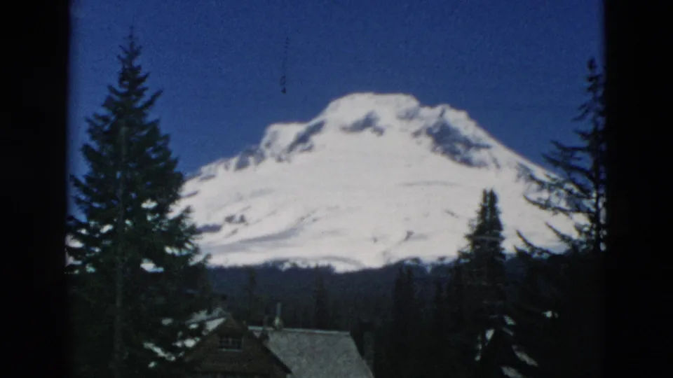 Archival 8mm view of a snow-covered mountain framed by tall evergreens, with part of a rustic building below.