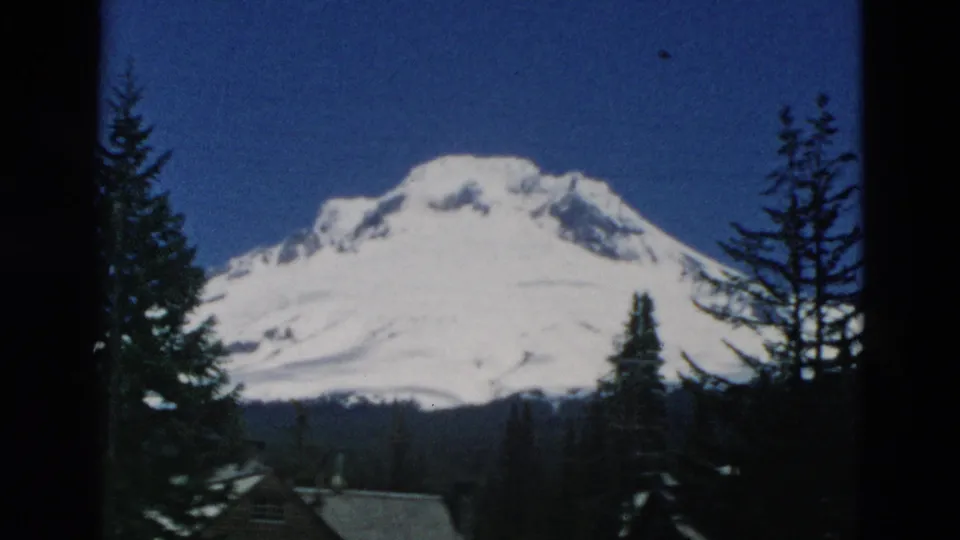 A snow-capped mountain is filmed through tall evergreens, with a low roofline and dark forested slope below.
