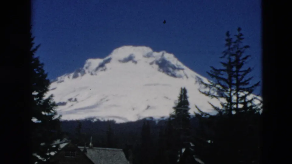 1950s 8mm frame of a snowy mountain viewed through evergreen silhouettes, with small rooflines low in the foreground.