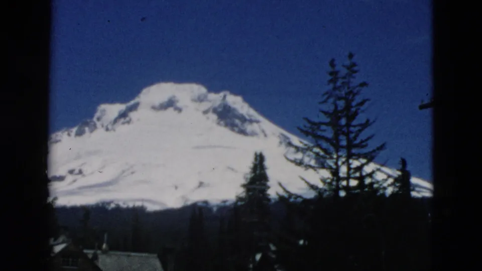 A snow-covered mountain rises behind pine-like trees, with the 8mm frame showing dark vertical borders and saturated blue sky.