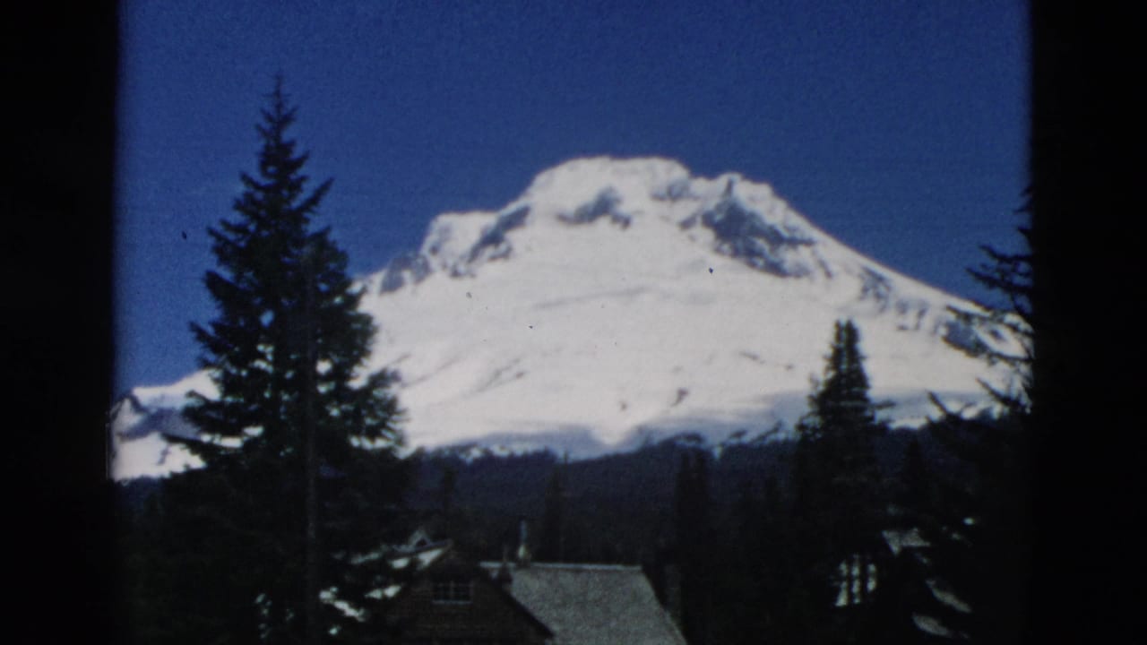 Snow-covered mountain behind evergreen trees and cabin roofs, framed by dark film borders.