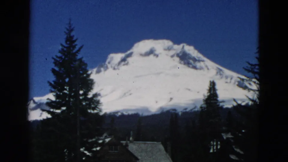 Likely Oregon 1950s 8mm view of a snowy mountain rising beyond evergreens and rustic cabin rooftops.