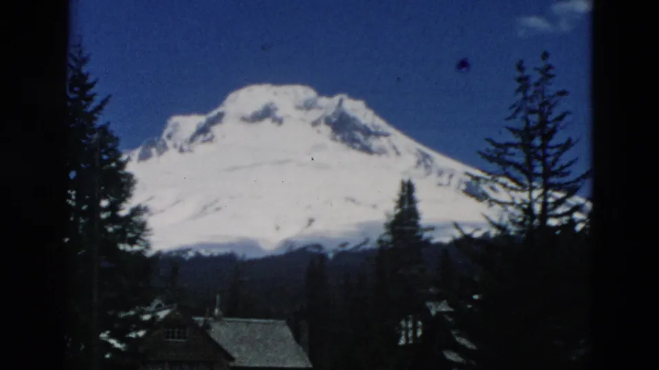 Likely Oregon mountain view on 8mm film, with a snow-covered peak behind evergreens and small cabin roofs.