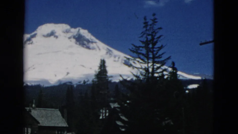 A vintage 8mm mountain view shows the snow-covered peak partly obscured by tall trees and cabin rooflines.