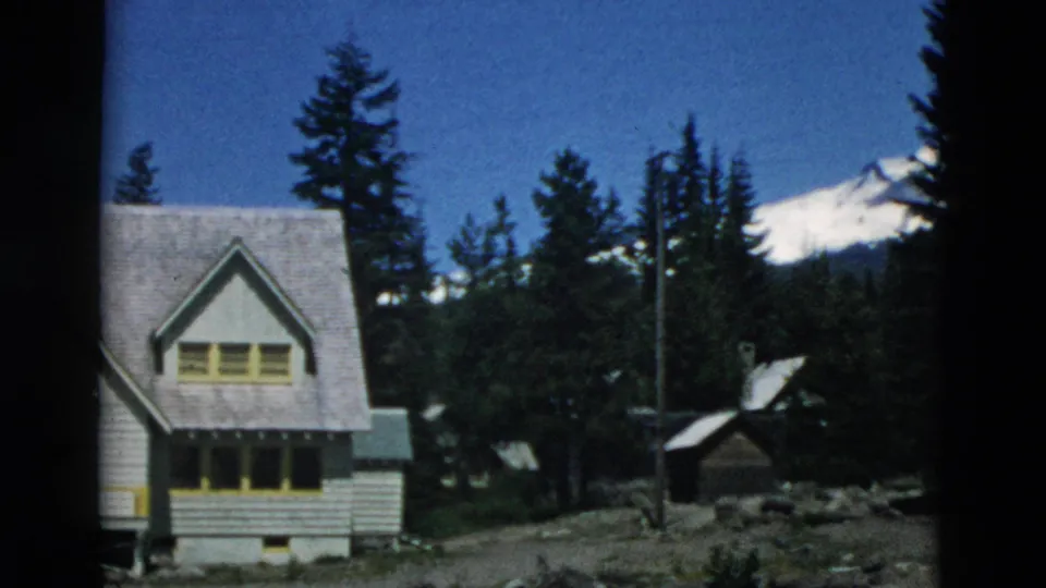 Likely Oregon 1950s 8mm frame showing a small house, evergreen trees, and a snowy mountain slope in the background.