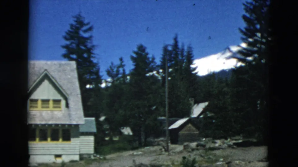 8mm frame of a maintained cabin, rustic outbuildings, tall evergreens, and a distant snow-capped mountain in archival home-movie footage.