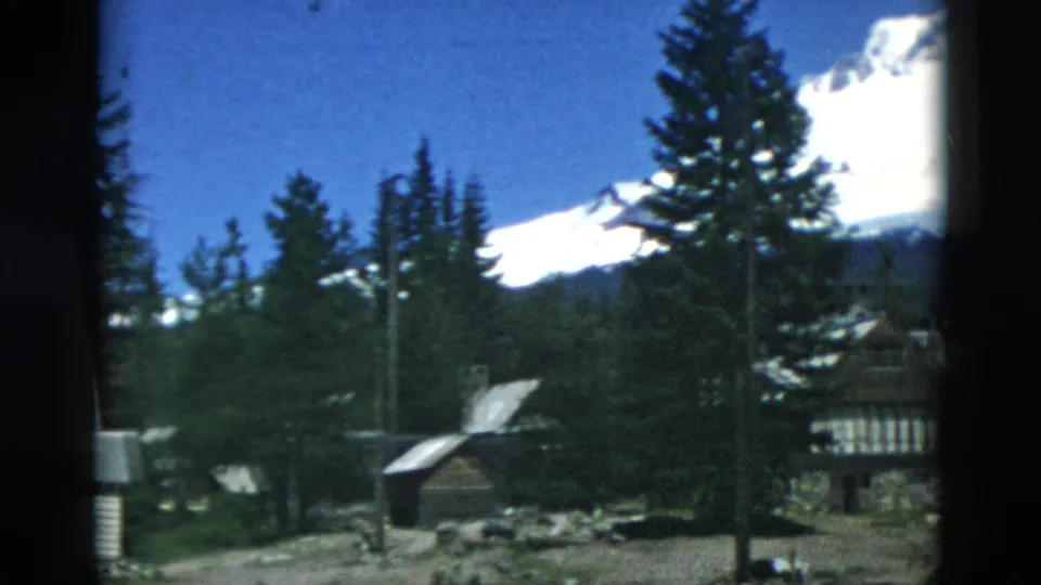 Archival 8mm view across rustic cabins and tall trees toward a snow-capped mountain under a clear blue sky.