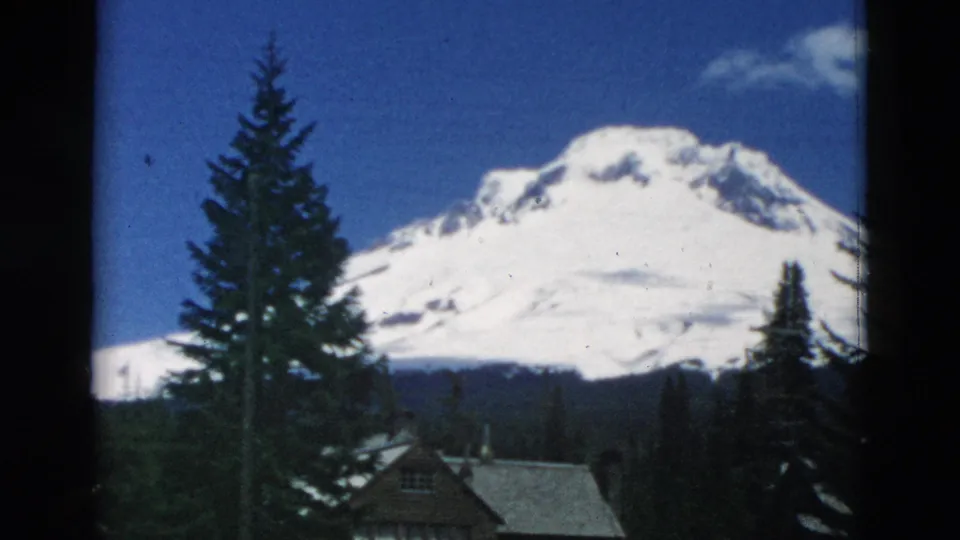 The camera view centers more on the snowy mountain, with dark evergreens, cabin roofs, and a utility pole in the foreground.