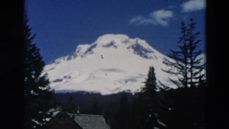 Archival home-movie frame of a snow-covered mountain above a forested foreground with cabin rooftops and tall conifers.