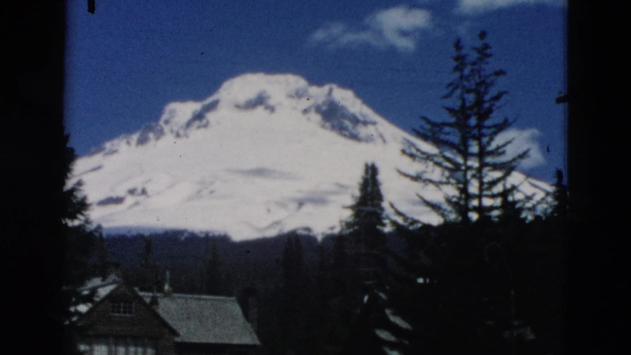 Snow-covered mountain behind evergreen trees with a cabin roof at the lower left.