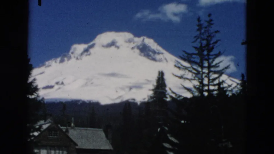 1958 8mm frame of a snowy mountain viewed past dark evergreens and a small cabin roofline under blue sky.