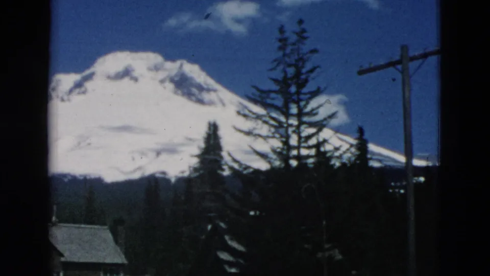 1958 8mm frame with a snow-capped mountain behind evergreens, a cabin roof, and a roadside utility pole.