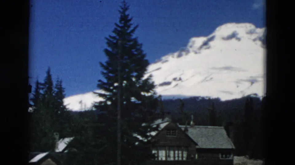 1958 8mm frame of a rustic cabin and evergreens beneath a snow-covered mountain, with blue sky above.