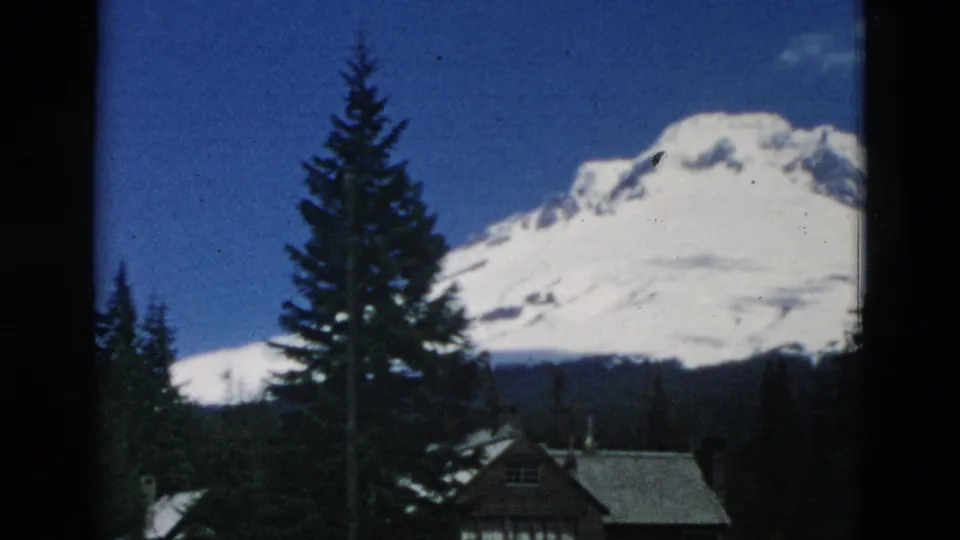 A snow-covered mountain rises behind dark evergreen trees, with part of a small cabin or lodge visible in archival 8mm footage.
