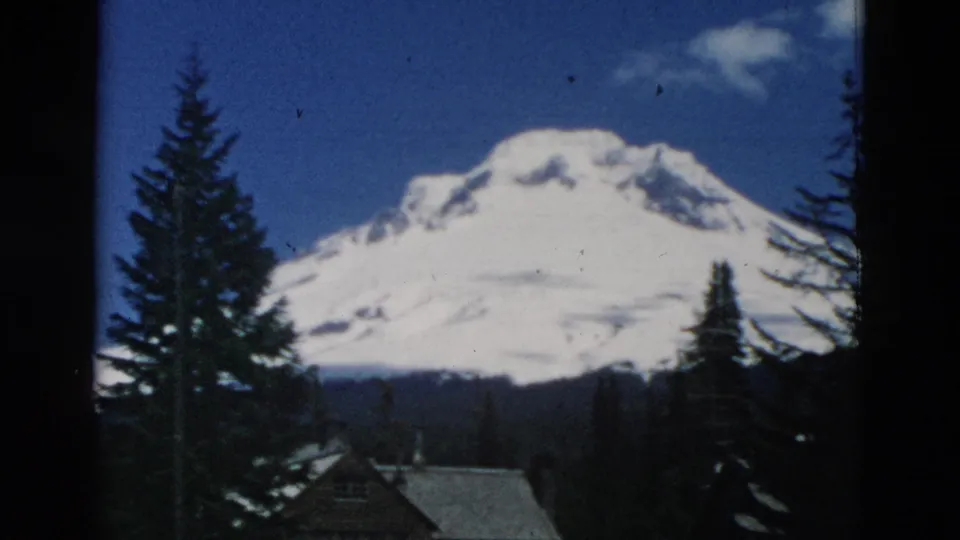 A broad snow-capped mountain is seen beyond evergreen trees and a low cabin or lodge roofline in soft 8mm color footage.