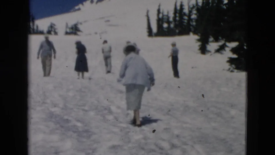Several adults in mid-century clothing walk and stand on a snowy slope, with trees and a mountain backdrop visible in the distance.