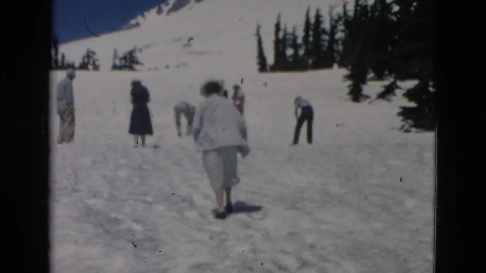A small group of adults crosses and explores a snowy open area beneath mountain slopes, with evergreen trees along the edge of the scene.