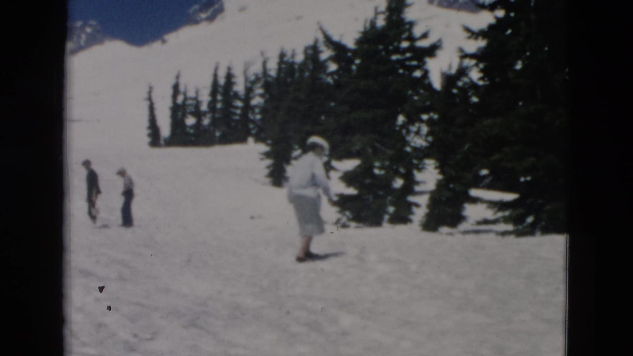 Woman in a large hat walks across snow near evergreen trees with two distant people at left.