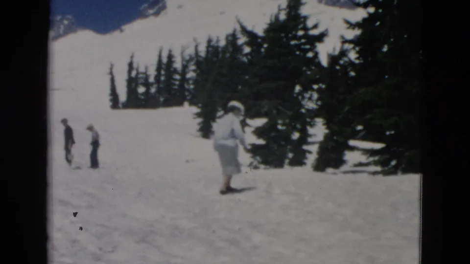 1950s 8mm frame of a woman in a light dress and large hat crossing a snowy slope near evergreens, with two people in the distance.