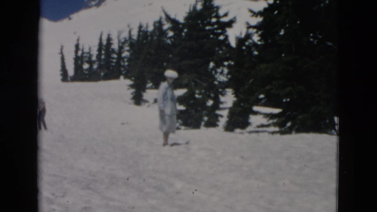 1950s home-movie frame showing a woman in a pale dress and broad hat on a snowy mountain slope near dark evergreens.