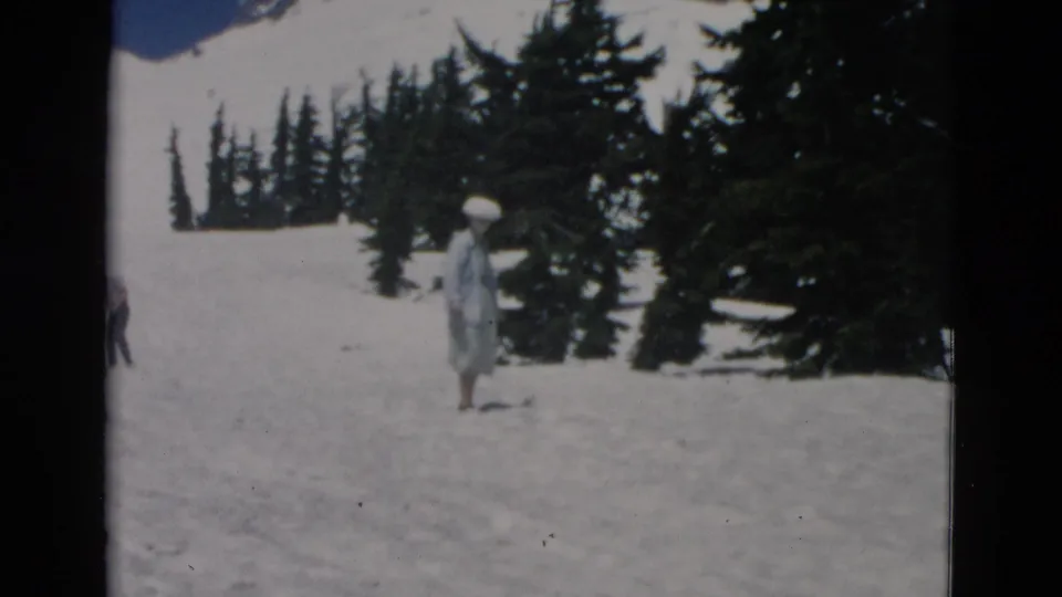 1950s home-movie frame showing a woman in a pale dress and broad hat on a snowy mountain slope near dark evergreens.