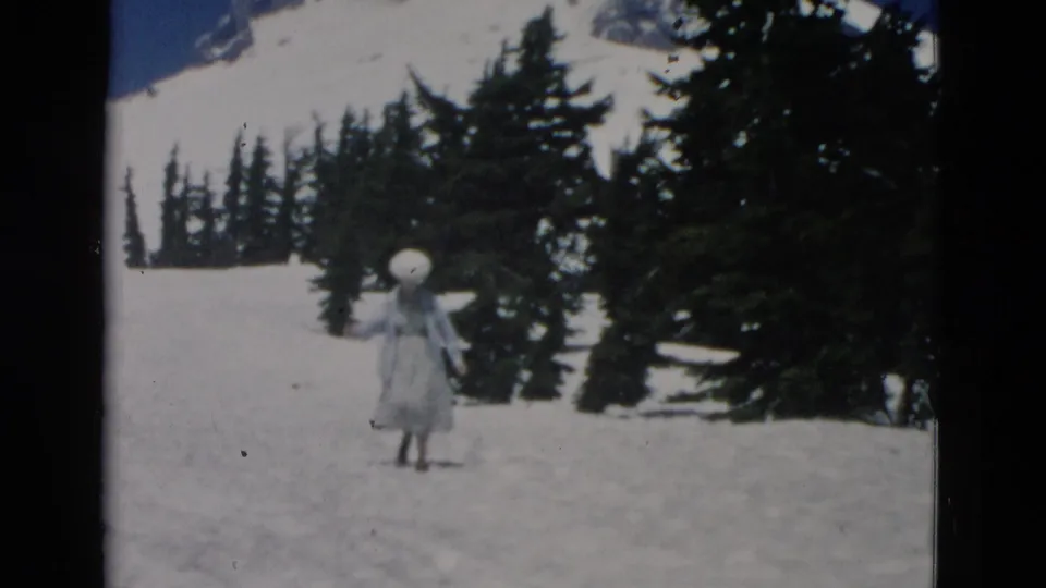 Small-gauge color frame of a woman in a light dress and large white hat walking through snow near a stand of evergreens.