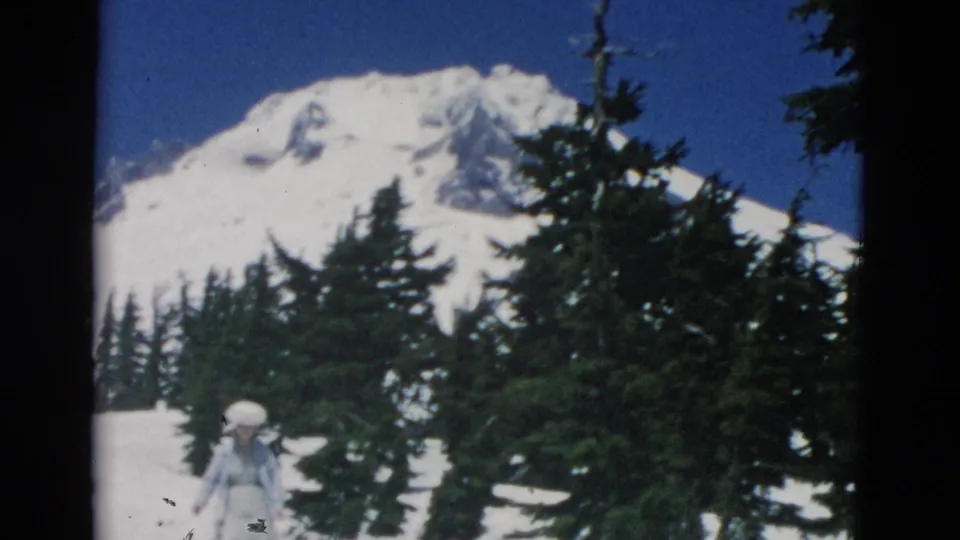 1950s 8mm frame with a snow-covered mountain peak, evergreen trees, and a woman in a large hat moving across the lower frame.