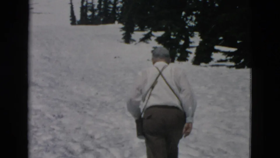 1958 Oregon 8mm frame showing an adult man in suspenders walking away uphill through snow near evergreens.