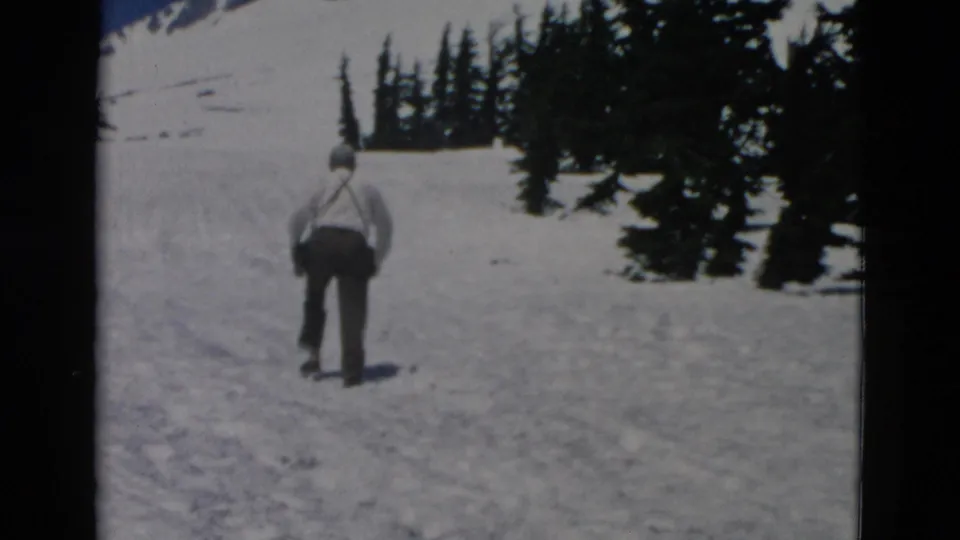 Wide 8mm view of a man receding uphill on a snowy mountain surface with dark evergreens beyond.