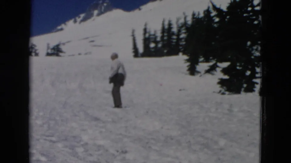 A man in light shirt and grey trousers stands on a bright snowy slope in 1958 home-movie footage, with trees and mountain snow behind him.