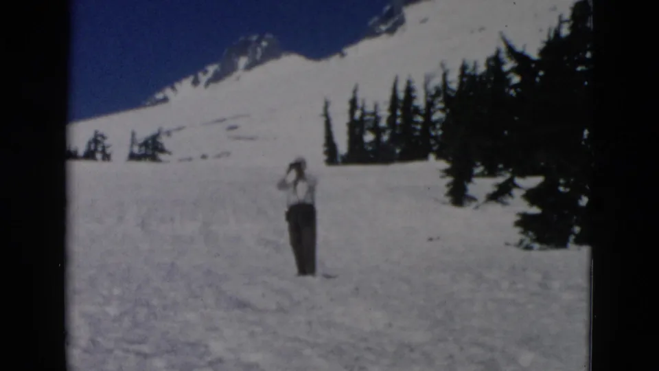 A man lifts binoculars toward his face on a bright mountain snowfield, framed by evergreen trees and distant snow-covered terrain.