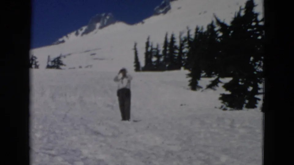 The man appears to look through binoculars while standing in snow, with dark evergreen trees and a bright mountain slope behind him.