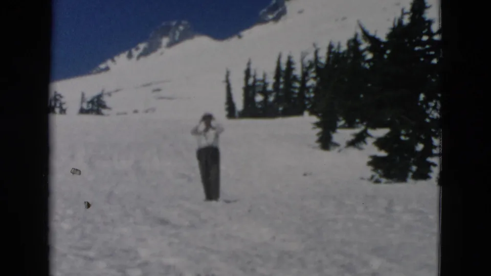 A man in dark pants stands on a snowfield below a snow-capped mountain, with evergreen trees under a blue sky.