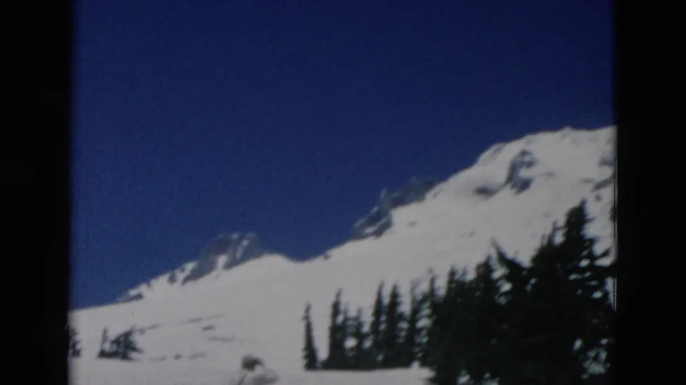 A soft 8mm frame shows the snow-covered mountain and dark evergreens, with the person only partly visible at the bottom.