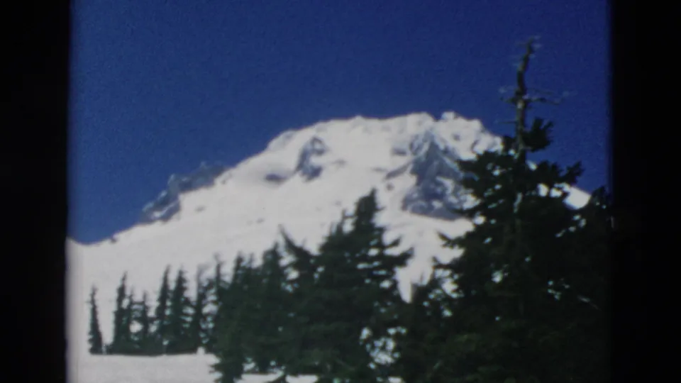 1958 Oregon 8mm frame showing a snowy mountain peak rising behind dark evergreen trees under a deep blue sky.