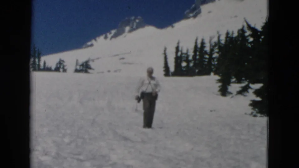 1958 Oregon 8mm frame showing an adult man walking across a snowy slope beneath a mountain ridge and evergreen trees.