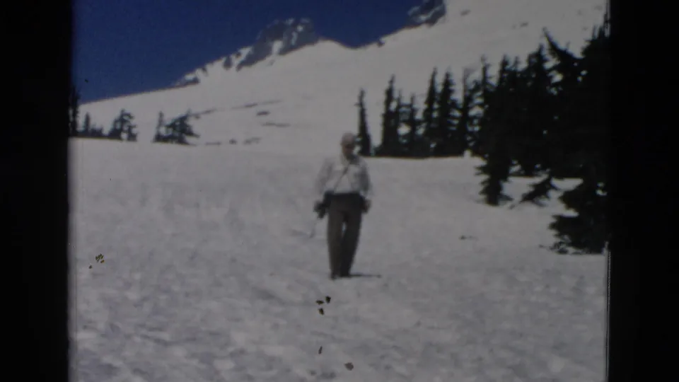 1958 Oregon 8mm frame of an adult man walking on snow, with evergreens and a mountain ridge in the background.
