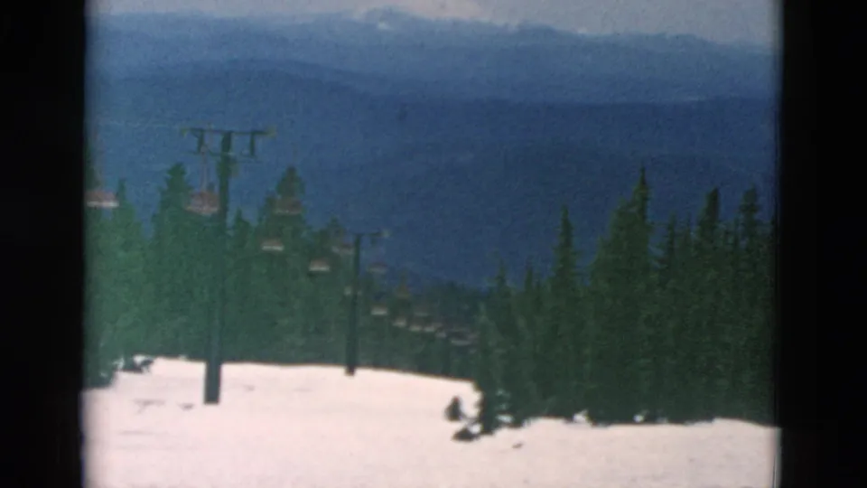 Reported Oregon ski-area view on 8mm film, showing snow, lift towers, dark evergreens, and layered mountain ridges beyond the slope.