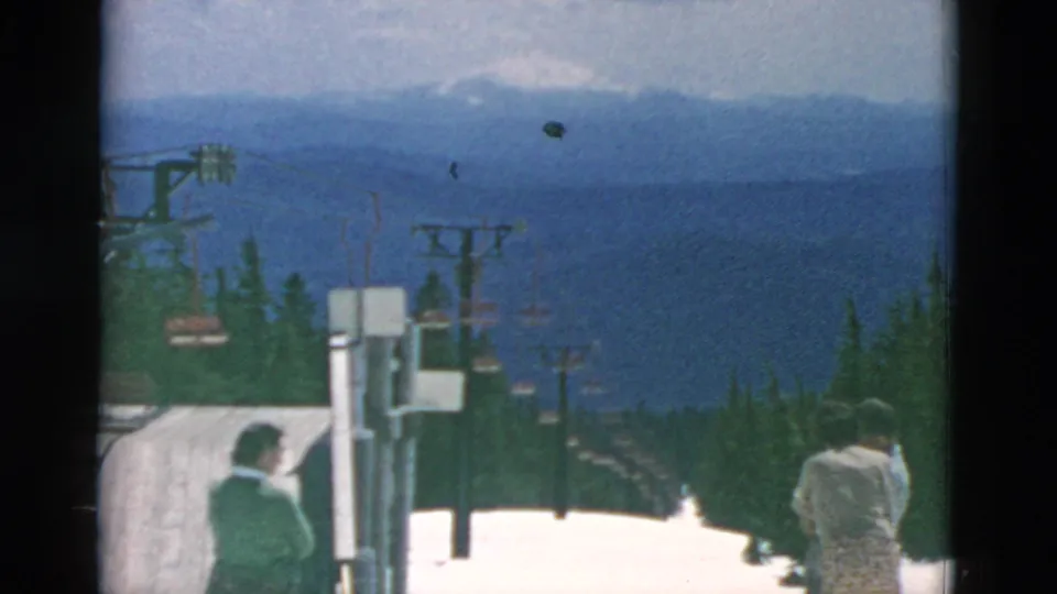 Two adults are visible near lift equipment at the snowy slope edge, looking toward a chairlift line, evergreens, and distant mountain ridges.