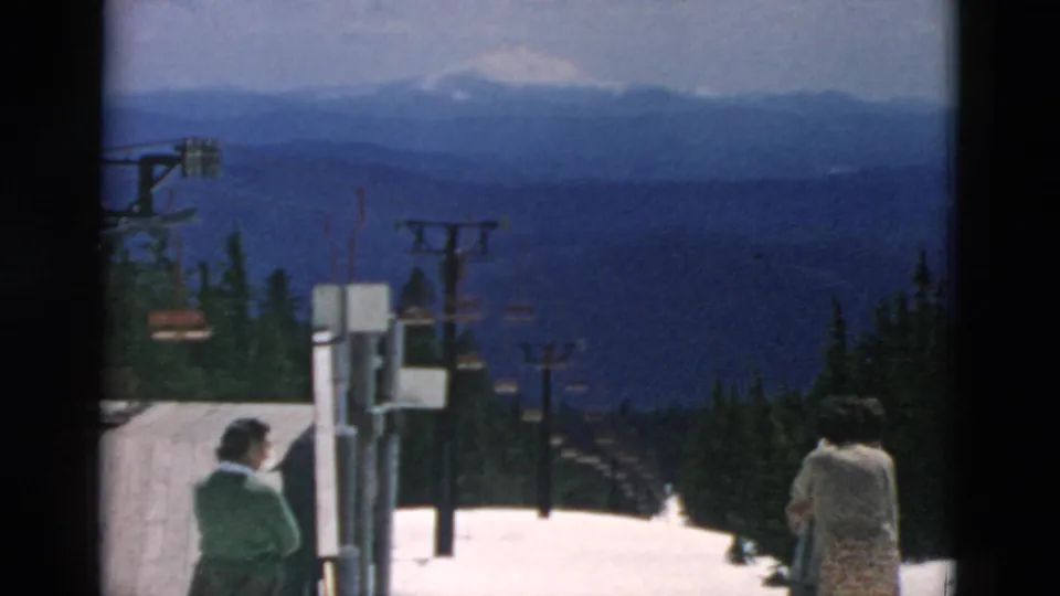 Adults stand partly turned away near the top of a reported Oregon ski slope, with lift structures, evergreens, and hazy mountain ridges ahead.